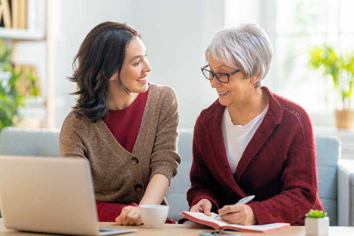 Adult daughter looking at older mother. Mother is looking at laptop screen and writing in notebook.