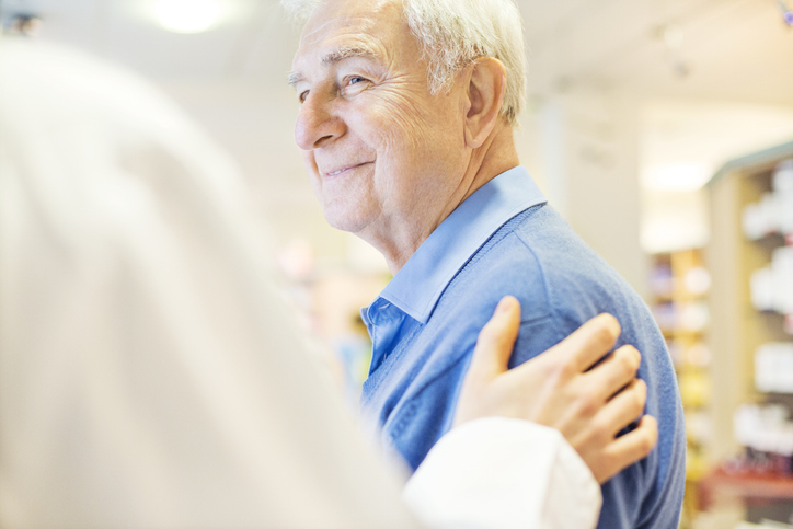 Cropped image of female pharmacist consoling senior man in store. Smiling male customer is looking at chemist. They are at brightly lit pharmacy.