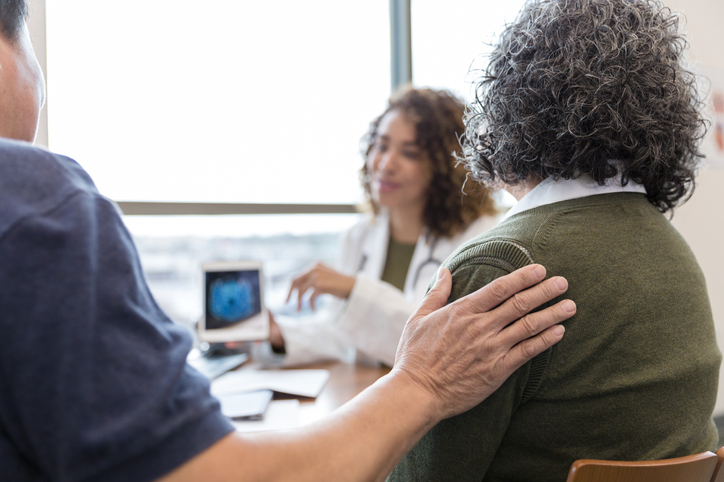 Close-up of a man's hand on his wife’s shoulder as he comforts her during a conversation with a female neurologist.