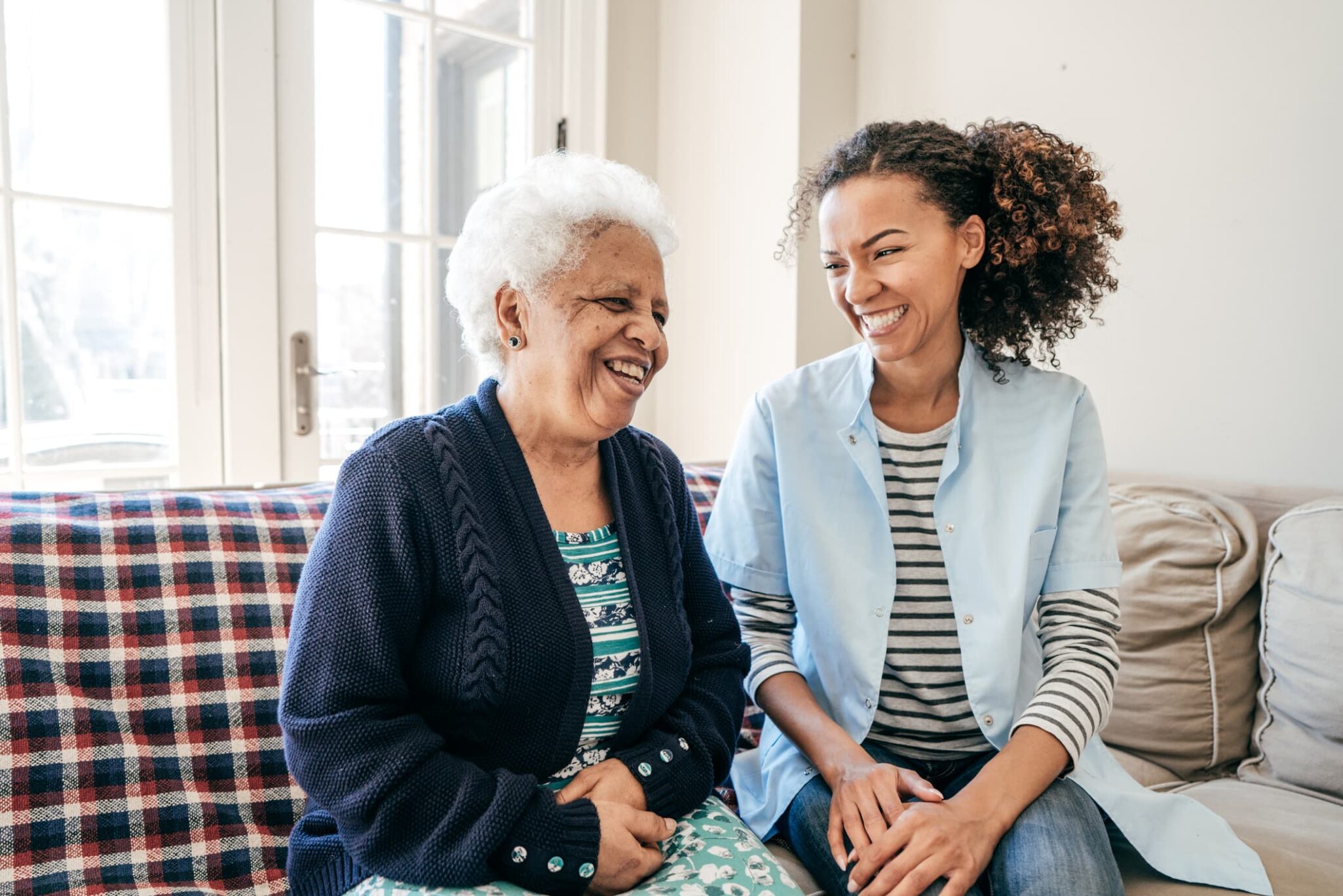 Elderly woman and young female caregiver sitting on couch and laughing.